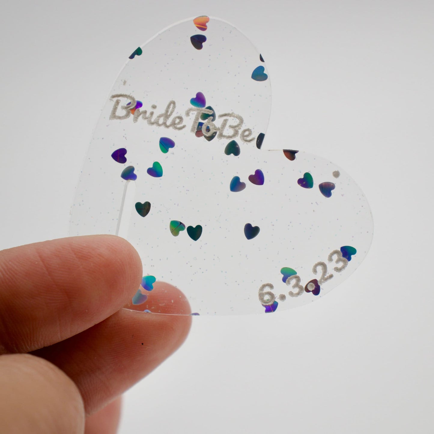 Close up of heart shaped drink tag with heart flakes acrylic to show the tiny hearts and soft shimmer. Painted on it says “bride to be 6.3.23” in white
