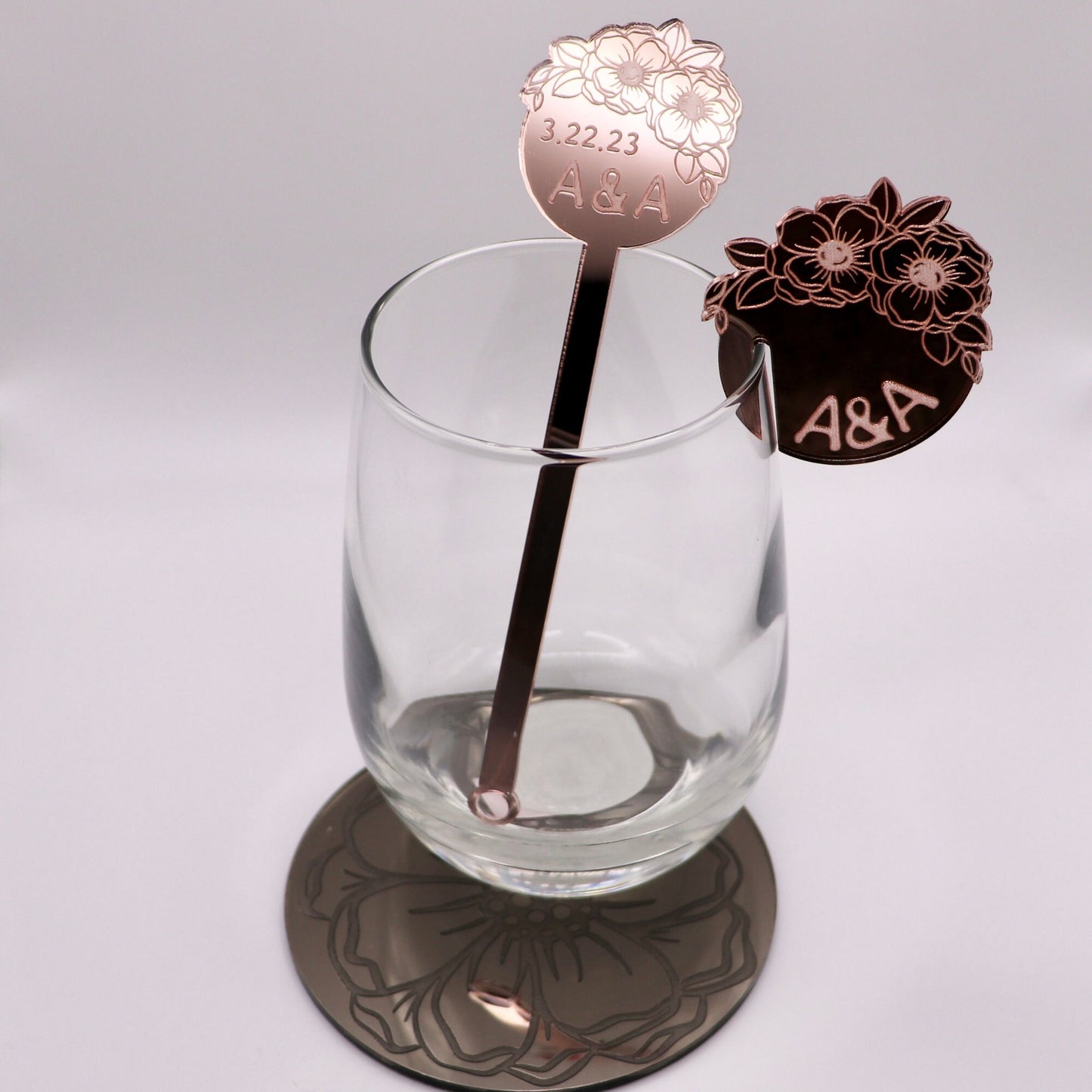 Rose gold mirrored drink stirrer in an empty wine glass with its matching drink tag. on top of a Bronze mirrored acrylic coaster. The items have Anemone flowers clustered together and engraved. Also engraved “3.22.23 A&A”