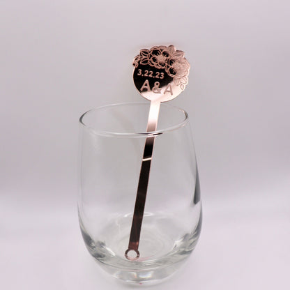 Rose gold mirrored drink stirrer in an empty wine glass on a white background. The drink stirrer has 2 Anemone flowers and leafs clustered together and engraved. Also engraved “3.22.23 A&A”