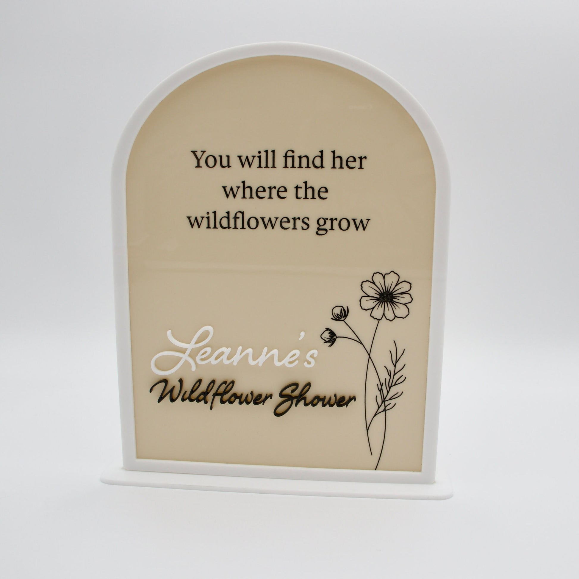 Arched acrylic sign in beige and white with black etched words. “You will find her where the wildflowers grow”Featuring an etched flower on the right. Anda second layer of acrylicas title “Leanne’s Wildflower shower”